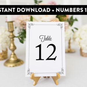 May include: White table number card with a black border and ornate corners. The card reads "Table 12" in black script font. The card is on a gold stand.