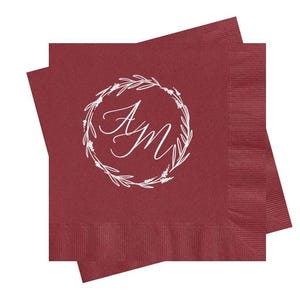 May include: Two square burgundy paper napkins stacked at an angle. Each napkin features a white printed design in the center consisting of a circular wreath made of thin leafy branches surrounding the stylized cursive letters AM in white ink.