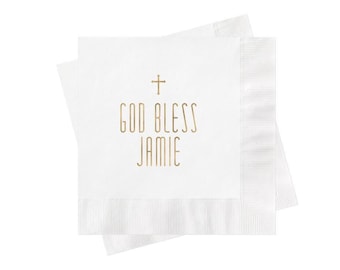 May include: Two square white paper napkins stacked on top of each other. The top napkin features a gold-colored cross symbol centered above the text "GOD BLESS JAMIE" printed in a clean, sans-serif gold font. The edges have a textured border.