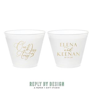 May include: Two clear plastic cups with gold lettering. One cup reads "One Day Away" and the other reads "ELENA and KEENAN 10.31.26". The cups are likely for a wedding or special event.