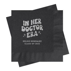 May include: Black paper napkins with silver text that reads "In Her Doctor Era" with stars and a flower design. The text "Kelsie Gonzalez Class of 2023" is also printed on the napkin.