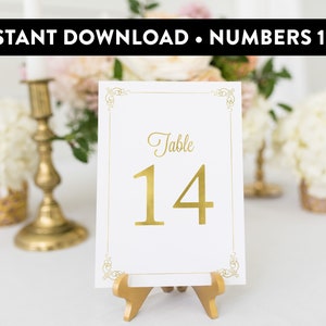 May include: Gold table number card with a white background and a gold frame. The card says "Table 14" in gold lettering. The card is on a gold stand.