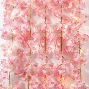 May include: A vertical arrangement of artificial cherry blossom vines in shades of light and dark pink. The delicate flowers are attached to thin, flexible stems and are interspersed with small, glowing warm white LED fairy lights.