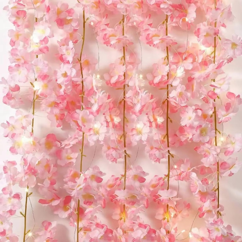 May include: A vertical arrangement of artificial cherry blossom vines in shades of light and dark pink. The delicate flowers are attached to thin, flexible stems and are interspersed with small, glowing warm white LED fairy lights.