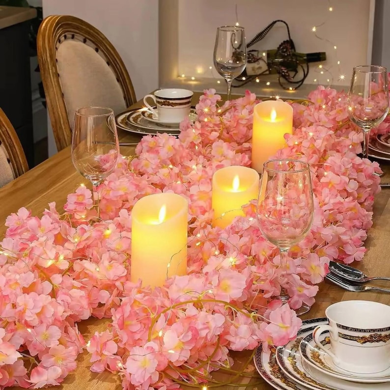 May include: A table centerpiece featuring a garland of pink artificial cherry blossoms intertwined with warm white fairy lights and three cylindrical LED pillar candles, arranged among clear glass wine glasses and white porcelain teacups.