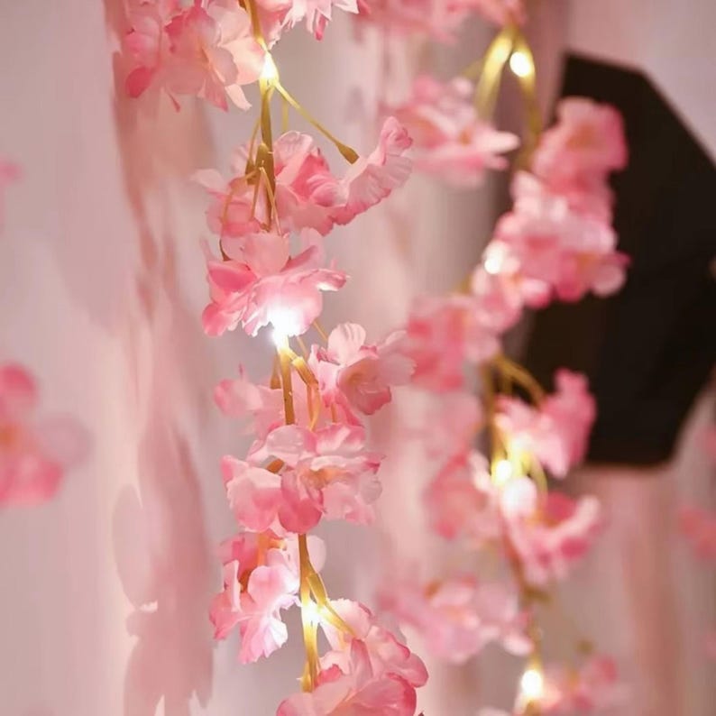 May include: A string of artificial cherry blossom flowers in shades of light and dark pink, featuring integrated small warm white LED lights nestled among the petals. The floral garland hangs vertically, creating a soft glow and delicate appearance.