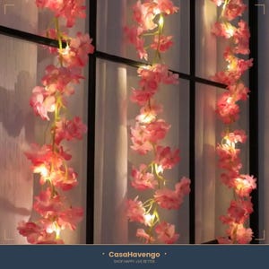 Könnte beinhalten: Eine Girlande aus künstlichen rosa Kirschblüten, die mit kleinen warmweißen LED-Lichterketten durchzogen ist. Die Blumengirlande hängt vertikal in drei Abschnitten und zeigt zarte Blütenblätter und grüne Stiele, die von den Lichtern beleuchtet werden.