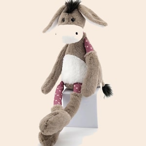 May include: A plush donkey toy with soft brown fur, featuring a white muzzle, a white belly patch, and long ears with dark tufts. The limbs are accented with a mauve fabric patterned with small white crosses, and the tail ends in a dark tuft.