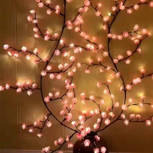 Op de afbeelding: Een decoratief arrangement van kunstmatige kersenbloesemtakken. De takken zijn donkerbruin, met talrijke roze bloesems en kleine lichtjes. Het arrangement staat in een bruine vaas, tegen een warmgekleurde achtergrond.