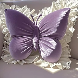 May include: A lavender butterfly-shaped pillow with a soft, velvety texture. The pillow has detailed wing patterns and a central body, resting on a white, lace-trimmed cushion. The design is decorative and whimsical.