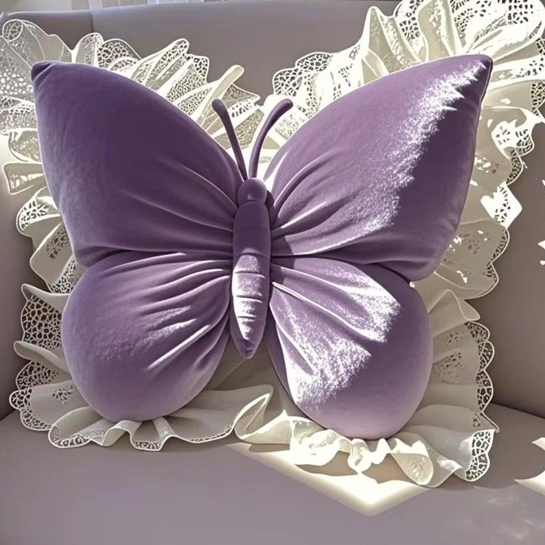 May include: A lavender butterfly-shaped pillow with a soft, velvety texture. The pillow has detailed wing patterns and a central body, resting on a white, lace-trimmed cushion. The design is decorative and whimsical.