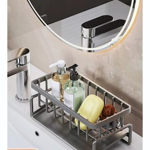 May include: A gray metal sink organizer holding soap, liquid soap dispensers, and a bar of soap. The organizer sits on a white sink, with a chrome faucet and a round mirror in the background.