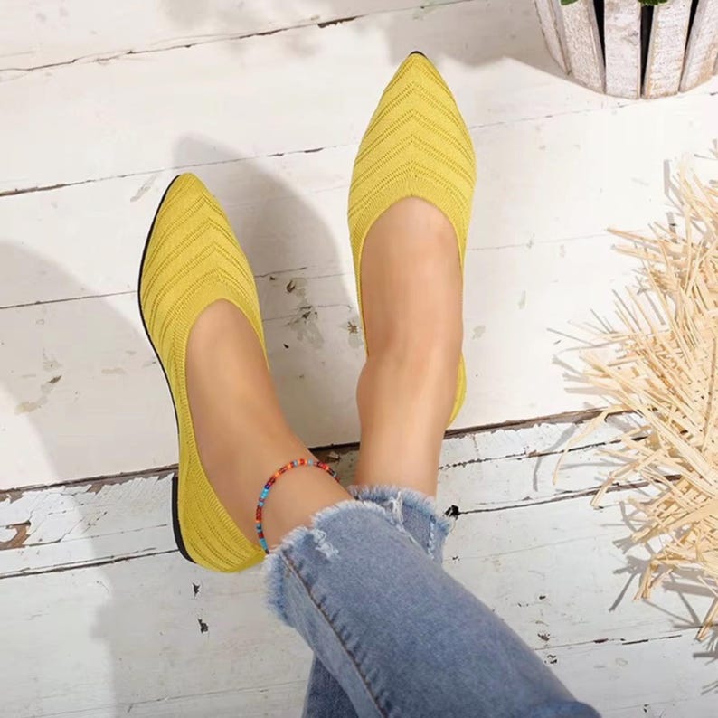 May include: A pair of yellow knit flats with a pointed toe. The shoes have a textured, striped pattern and a black sole. The image shows the shoes being worn with blue jeans and a colorful ankle bracelet. The background is a white wooden surface.