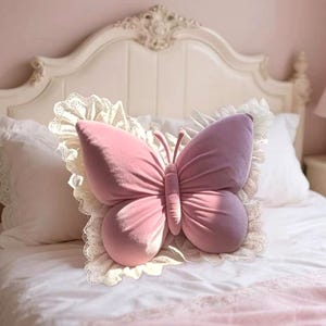 May include: A pink butterfly-shaped pillow with a velvet texture rests on a bed. The pillow is surrounded by a white lace trim. The bed has white linens and pillows, and a decorative white headboard.