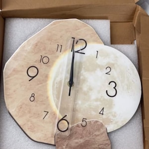Puede incluir: Un reloj de pared decorativo con un diseño único. La esfera del reloj presenta una forma beige similar a la piedra con números y manecillas negras. El reloj tiene un diseño similar a la luna con un color beige claro. El reloj está embalado en una caja de cartón.