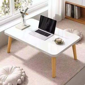 Op de afbeelding: Een witte rechthoekige lage tafel met vier lichtgekleurde houten poten. Op het tafelblad staan een open zilveren laptop, een klein wit boek en een keramieken kopje op een schoteltje. De tafel staat op een ondergrond van lichtgekleurde stof.