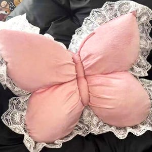 May include: A pink butterfly-shaped pillow with a white lace trim. The pillow has a soft, plush texture and is designed to resemble a butterfly with its wings spread. The lace trim adds a delicate touch to the overall design.