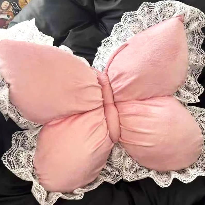 May include: A pink butterfly-shaped pillow with a white lace trim. The pillow has a soft, plush texture and is designed to resemble a butterfly with its wings spread. The lace trim adds a delicate touch to the overall design.