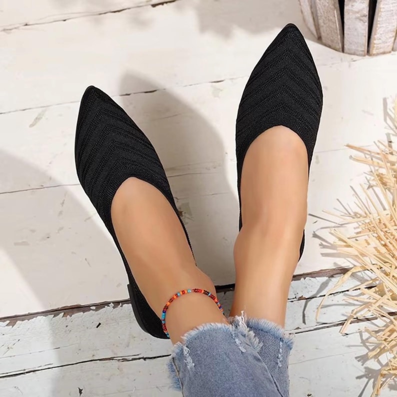 May include: Black knit flats with a pointed toe. The shoes have a low-cut vamp and a textured knit pattern. A colorful beaded bracelet is worn on the ankle. The shoes are shown on a white wooden surface.