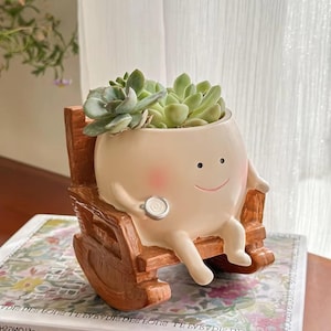 May include: A whimsical, beige ceramic planter shaped like a smiling character seated in a wooden rocking chair. The planter holds several green succulents. The character has rosy cheeks and is holding a small, round object.