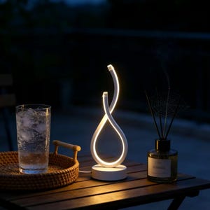 May include: A white, modern table lamp with a unique, curved design. The lamp has a teardrop shape with a glowing interior, and sits on a round base. A glass of water with ice, a woven tray, and a diffuser bottle are also on the table.