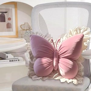 May include: A pink butterfly-shaped pillow with a soft, velvety texture. The pillow is centered on a white chair and is surrounded by a cream-colored lace trim. The butterfly has detailed antennae and wing shapes, creating a decorative accent.