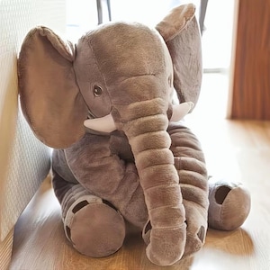 May include: A plush elephant toy made of soft, grey fabric with large ears, a long trunk, and two small white tusks. The stuffed animal is seated in an upright position, featuring detailed stitching on the trunk and feet, with dark eyes.