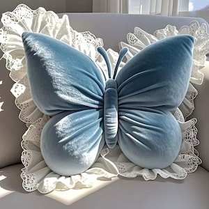 May include: A light blue butterfly-shaped pillow with a soft, velvety texture. The pillow has a detailed design, including antennae and wing shapes. The butterfly is set against a white, lace-trimmed background, creating a decorative accent.