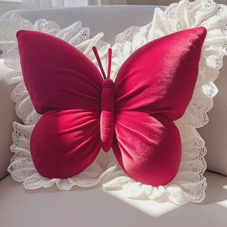 May include: A plush, crimson butterfly pillow with a soft, velvety texture. The butterfly is centered on a white, lace-trimmed pillow, creating a delicate contrast. The pillow's wings are spread wide, and the antennae are visible.