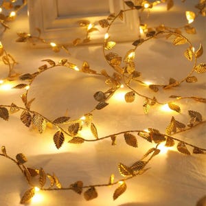 May include: Gold-colored leaf string lights. The lights are arranged in a garland shape, with small, warm white lights interspersed among the metallic leaves. The leaves have a detailed, textured appearance, and the overall effect is elegant and decorative.