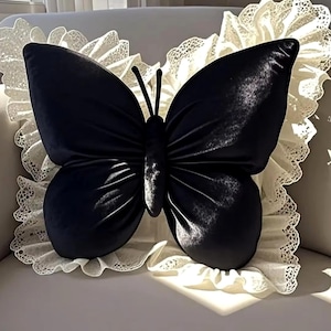 May include: A black butterfly-shaped pillow with a velvet texture. The pillow is centered on a white, ruffled lace trim. The butterfly has detailed wings and antennae. The pillow is a decorative home accent.