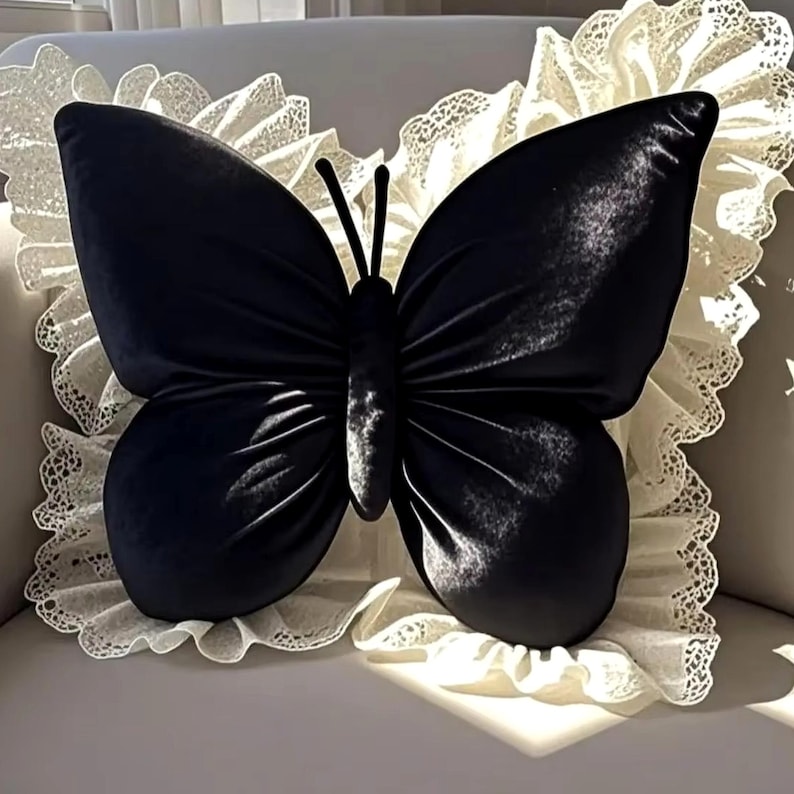 May include: A black butterfly-shaped pillow with a velvet texture. The pillow is centered on a white, ruffled lace trim. The butterfly has detailed wings and antennae. The pillow is a decorative home accent.