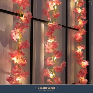 May include: A string of artificial pink cherry blossom flowers intertwined with small warm white LED fairy lights. The floral garland hangs vertically in three sections, featuring delicate petals and green stems illuminated by the glowing lights.