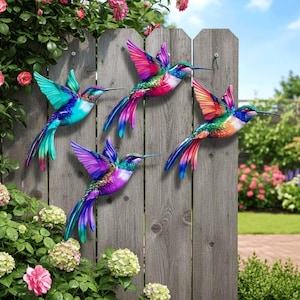 May include: Four metal hummingbird wall sculptures in vibrant colours including teal, magenta, purple, and orange. Each bird is depicted in a dynamic flying pose with outstretched wings and long tail feathers, mounted on a vertical wooden fence panel.