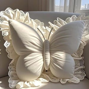 May include: A plush, off-white butterfly-shaped pillow with detailed wing and body stitching. The pillow is set against a backdrop of a white lace-trimmed pillow, creating a soft, elegant aesthetic.