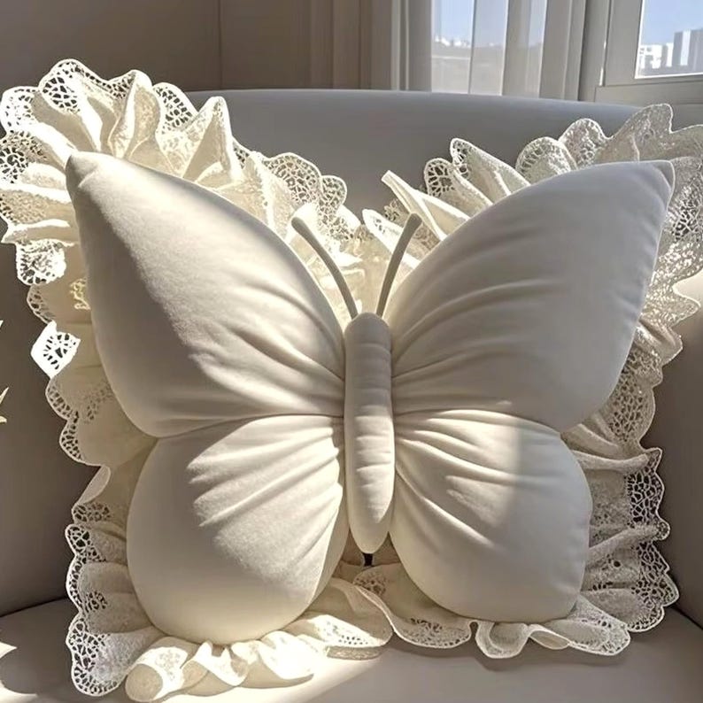 May include: A plush, off-white butterfly-shaped pillow with detailed wing and body stitching. The pillow is set against a backdrop of a white lace-trimmed pillow, creating a soft, elegant aesthetic.