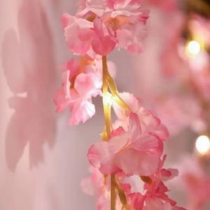 May include: A string of artificial cherry blossom flowers in shades of light and dark pink, featuring integrated warm white LED lights along the stems. The delicate petals are layered and soft, creating a glowing floral display with soft shadows.
