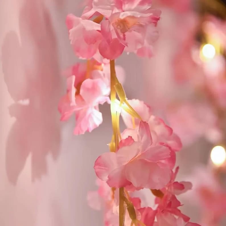 May include: A string of artificial cherry blossom flowers in shades of light and dark pink, featuring integrated warm white LED lights along the stems. The delicate petals are layered and soft, creating a glowing floral display with soft shadows.