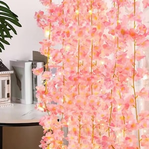 May include: A hanging string of artificial pink cherry blossom flowers intertwined with thin, glowing LED fairy lights. The delicate pink petals are arranged along slender, flexible stems, creating a cascading floral display with warm light.