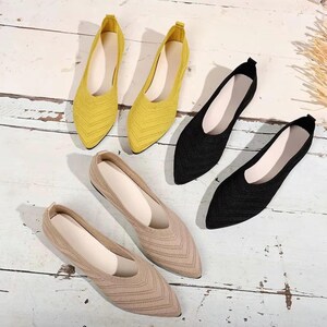 May include: A collection of pointed-toe flats in yellow, black, and beige. The shoes have a knitted texture and a low profile. The footwear is arranged on a light-colored wooden surface, showcasing the variety of colors and styles.