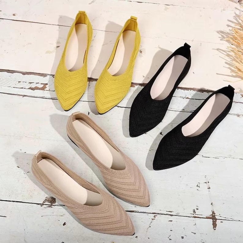 May include: A collection of pointed-toe flats in yellow, black, and beige. The shoes have a knitted texture and a low profile. The footwear is arranged on a light-colored wooden surface, showcasing the variety of colors and styles.