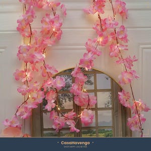 May include: A decorative string of artificial pink cherry blossom flowers intertwined with small warm white LED fairy lights. The floral garland features multiple clusters of soft pink petals on thin brown stems, creating a cascading effect.