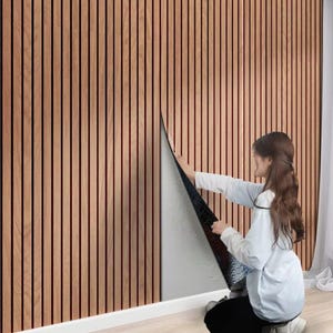 May include: A wall covering featuring vertical light brown wood slats with a natural grain pattern. The paneling has a black backing and is shown being applied to a wall, revealing a dark adhesive layer on the underside of the flexible material.