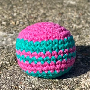 May include: A handmade, round crochet ball in vibrant pink and teal. The ball features alternating bands of bright pink and teal yarn, creating a striped pattern. The texture of the crochet stitches is clearly visible, giving it a handcrafted look.