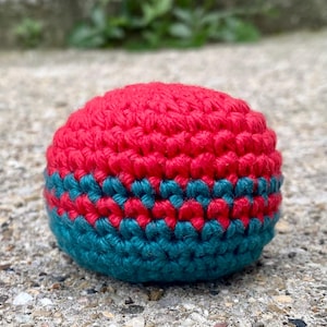 May include: A spherical, hand-crocheted ball featuring a vibrant red top section and a deep teal bottom section. The two colors are separated by a distinct horizontal band where the red and teal yarn stitches alternate in a repeating pattern.