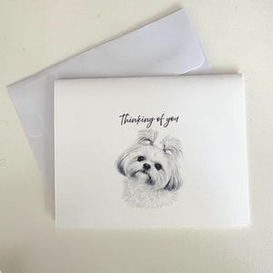 May include: A white greeting card with a pencil drawing of a Shih Tzu dog and the words "Thinking of you" in cursive. The card is accompanied by a white envelope, all set against a white background.