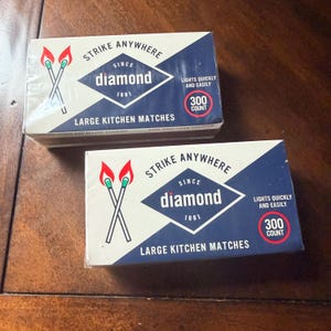 May include: Two rectangular boxes of Diamond brand large kitchen matches. Each box features a blue and white design with the text Strike Anywhere, Since 1881, Diamond, Lights Quickly and Easily, and 300 Count. Two lit match illustrations appear.