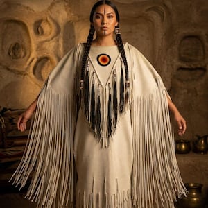 May include: A cream-colored dress with long fringe sleeves and hem. The dress features a geometric design with a circular emblem at the neckline. The model has long braids and traditional face paint. The background is a cave setting.