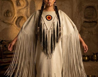 May include: A cream-colored dress with long fringe sleeves and hem. The dress features a geometric design with a circular emblem at the neckline. The model has long braids and traditional face paint. The background is a cave setting.