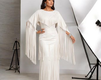 May include: A floor-length off-white dress featuring a V-shaped fringe overlay on the bodice and long, flowing fringe sleeves. The garment has a fitted waist and a straight skirt that is also adorned with long fringe detailing at the hemline.